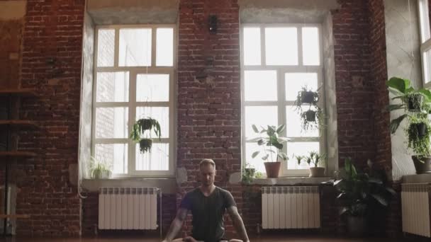 蓮の中でヨガを練習する男性は屋内でポーズをとります 木製の床と大きな窓のあるスタジオでヨガをしている若者 ヨガの概念 コピースペースでカメラがスライドダウン — ストック動画