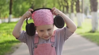 Kulaklık takan, müzik keyfi gülümseyen küçük kızın yakın portresi. Açık havada kablosuz kulaklıklı pembe güneş elbisesi giymiş çocuk