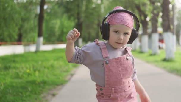 ワイヤレスヘッドフォンを着用し 音楽を聴き カメラを見て 踊る幸せな小さな女の子 ミディアムショット 屋外ピンクのサンドレスでかなり白人の小さな女の子 — ストック動画