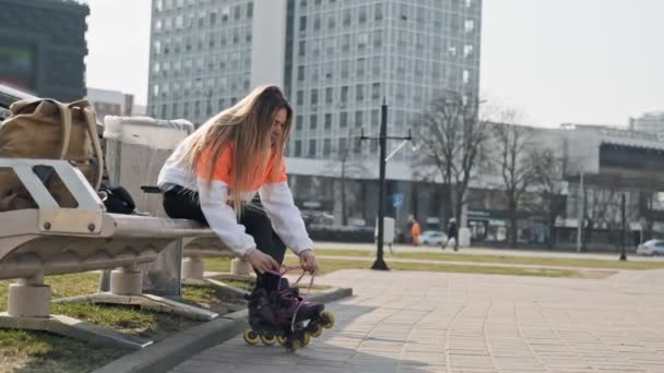 坐在街道背景的木凳上 年轻女子穿上滚筒的侧视图 美丽的年轻女孩把溜冰鞋在溜冰公园与复制空间 — 图库视频影像