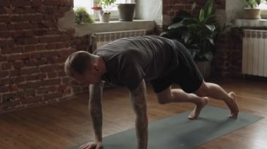 Genç adam içeride yoga yapıyor. Erkek yavaş çekimde kapalı siyah mat üzerinde bir asana yapıyor. Tam uzunlukta çekim. Adam meditasyon