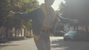 Genç kız yazın güneş ışınları eşliğinde şehrin sokaklarında modern hip hop dansı yapıyor, kıvrılıp dönüyor. Ağır çekimde dans eden kadının orta boy fotoğrafı. At kuyruklu kot giyen bir kadın.
