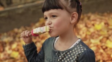 Mutlu kız şeker yiyor ve sonbahar parkında gülümsüyor. Dışarıda yüz yüze mutluluk. Görünümü kapat