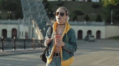 Gün batımında şehrin sokaklarında saçı örgülü genç bir kadın yürüyor. Güneş gözlüklü güzel kız şehir yürüyüşünde kahve içiyor. Orta boy. Yaşam tarzı kavramı