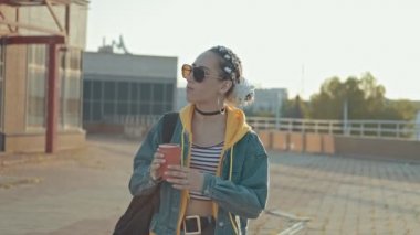 Kot pantolonhipster kız, örgülü kapüşonlu açık havada. Çekici genç kadın çay veya kahve fincan güneş gözlüğü güneşli şehirde yürüyüş. Orta atış
