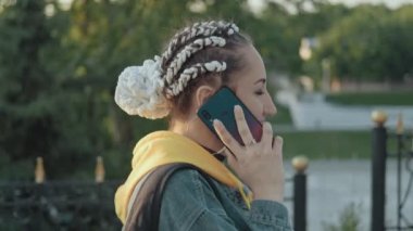 Sokakta yürüyen ve telefonla konuşan genç bir kadın. Takip kamerasıyla yakın çekim. At kuyruklu kadın kot pantolon giyiyor..