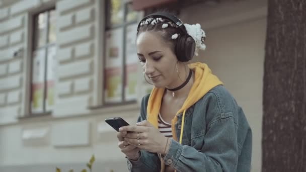 音楽を聴いて楽しい若い女性はイヤホンを着用します ジーンズをはいている女性携帯電話を使用してスタイリッシュな女の子のテキストメッセージ ソーシャルネットワークの概念 — ストック動画
