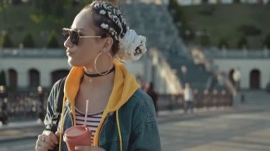 Çekici genç kadın çay veya kahve fincan güneş gözlüğü güneşli şehirde yürüyüş. Kot pantolonhipster kız, örgülü kapüşonlu açık havada. Orta atış