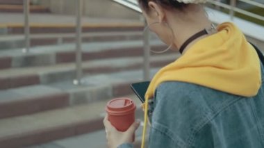 Sarı kapüşonlu çekici genç kadının arka görünümü yürüyüş, telefon kullanarak metin mesajlaşma ve kahve veya çay içme. İzleme kamerası. Yakın çekim görünüm