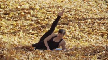 Atletizm kadın açık havada sarı yaprakları ile çayır bacaklar germe. Yavaş çekimde sonbahar parkında egzersiz güzel uygun kadın tam uzunlukta çekim. Spor konsepti