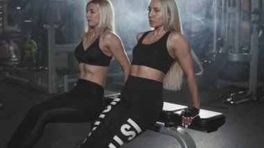 Fitness genç kadınlar gönderme yapıyor ups yavaş koyu spor salonunda. Atletik kızlar triceps bankta egzersiz. İki Sportif sarışın kız şınav gerçekleştirme
