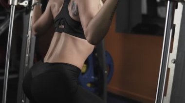 Sarışın atletik kadın yavaş hareket bacaklar için güç egzersizleri gerçekleştirir. Simülatörü Smith. Ağırlığı ile squatting. Orta atış. Karanlık spor salonunda fitness için egzersiz. Kız simülatörü egzersizleri yapıyor