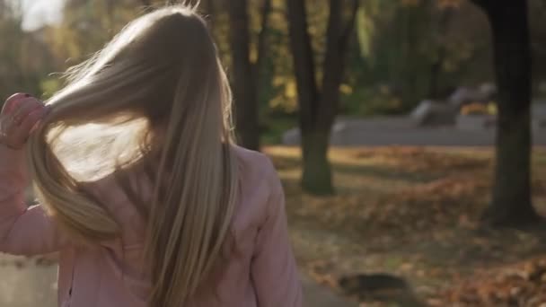笑顔の女の子は振り向き スローモーションで秋の公園のカメラを見て笑顔 夕暮れ時の魅力的なブロンド女性の肖像 太陽が輝いている ミディアムショット — ストック動画