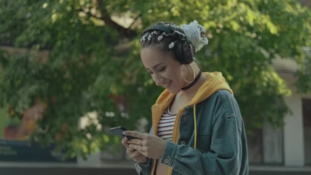 有吸引力的年轻女子在城市步行时使用智能手机在无线耳机听音乐 微笑的女孩与辫子的肖像 慢动作中镜头 — 图库视频影像