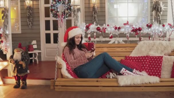 サンタの帽子をかぶった魅力的なブルネットの女性は 電話とコーヒーを飲んでクリスマスで誰かを迎えます ベンチに座っているクリスマスツリーの近くに電話でかわいい女の子のテキストメッセージ フルレングスショット — ストック動画