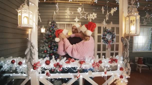 クリスマスツリーとポーチで抱き合うサンタクロースの帽子に身を包んだ母 小さな娘の若い幸せな家族 スローモーションで家族 Xmasコンセプト — ストック動画