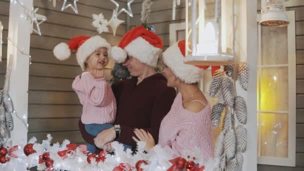クリスマスツリーとポーチで抱き合うサンタクロースの帽子に身を包んだ母 父と小さな娘のかわいい家族 スローモーションで家族の概念 — ストック動画