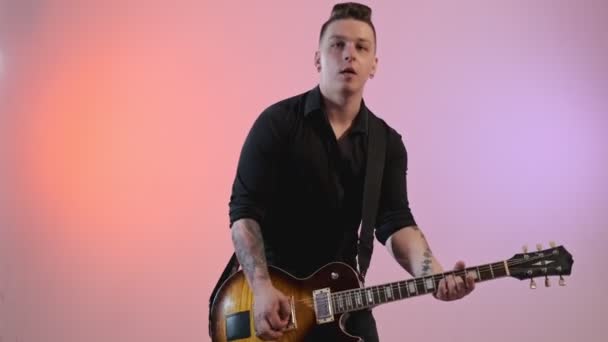 셔츠를 잘생긴 남자가 배경에 빨간색과 보라색 조명으로 일렉트릭 기타를 연주합니다 — 비디오