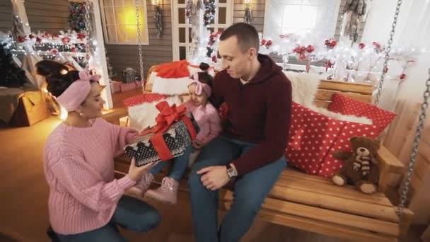 クリスマスのブランコで小さな娘に贈り物をする母 ポーチのクリスマスツリーの近くでブランコに座っている家族 スローモーションで家族とクリスマスの概念 — ストック動画