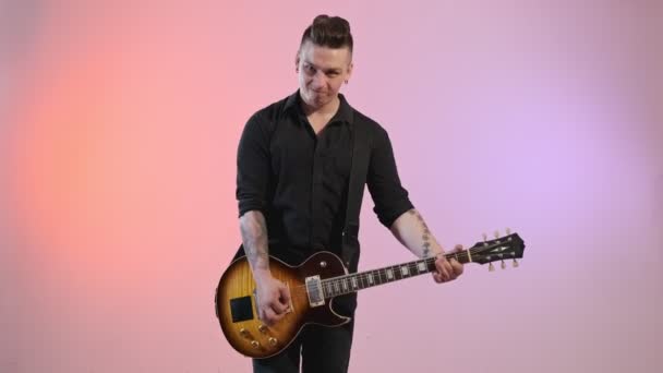 ギターで若い入れ墨の男のミディアムショット カラフルな背景を持つ屋内でエレキギターを演奏黒シャツの男性ギタリスト — ストック動画