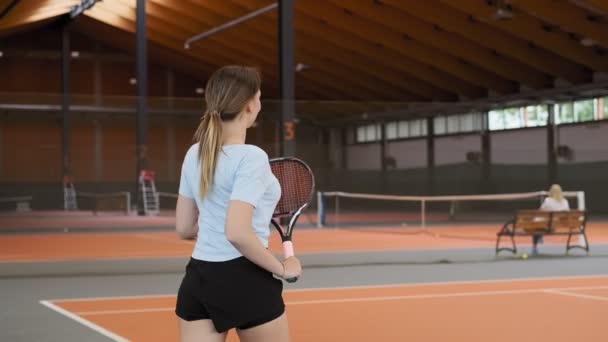 年轻女子在橙色网球场上行走 手里拿着球拍的侧视图 有吸引力的女选手在室内的硬地球场上行走的中拍 — 图库视频影像