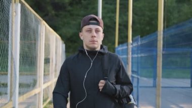 Odaklanmış genç adam güneşli sabah eğitim için yürüyüş. Kulaklık ve sırt çantası ile kap sporcu ön görünümü. Orta, atış takip edin. Spor konsepti