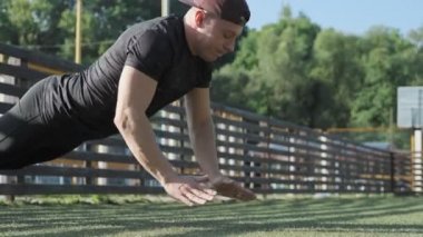 Yakışıklı adam stadyumda el çırparak şınav yapıyor. Güneşli bir günde spor egzersizleri yapan şapkalı güçlü adam. Egzersiz