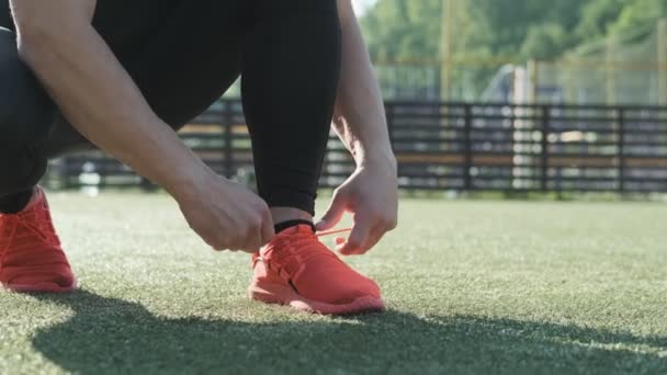 年轻运动员在绿色游乐场的鞋的特写镜头 运动员在户外训练前把鞋带绑在橙色运动鞋上 低视图 体育概念 — 图库视频影像