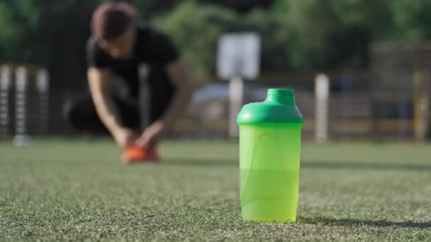 运动瓶水的特写镜头站在操场上 运动员在模糊的背景上绑鞋 男人拿瓶子运动与健康生活方式理念 — 图库视频影像