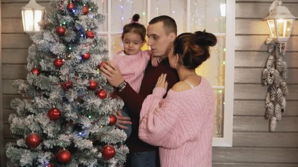 スローモーションでポーチで一緒にクリスマスと新年の木を飾るプルオーバーで母 父と小さな娘の幸せな家族 ミディアムショット — ストック動画