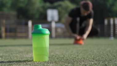 Oyun alanında, bulanık arka planda ayakkabı bağlayan sporcuile bir şişe su. Spor konsepti. Yeşil şişenin yakın çekim görünümü