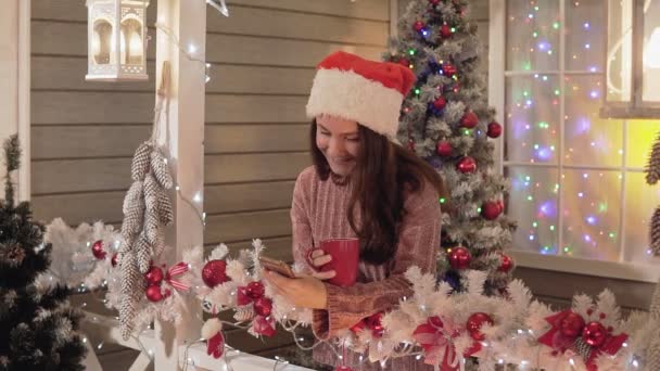 年轻的微笑的女人在圣诞老人的帽子微笑 并发送问候短信使用智能手机在圣诞树背景 美丽的女人拿着手机和红茶 右侧复制空间 — 图库视频影像