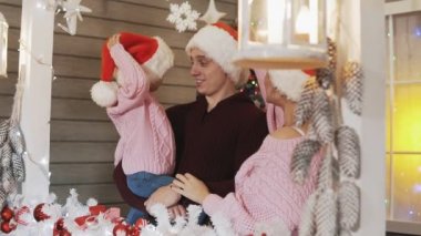 Anne, baba ve küçük kızı genç mutlu aile pullovers ve arka plan sundurma, Noel ve yılbaşı ağacının, sarılma Santa Claus şapka giymiş. Aile, Xmas kavram yavaş hareket