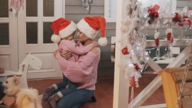 Genç anne ve kızı pembe pullovers ve kırmızı şapka Noel'de sarılma. Mutlu anne Noel verandada onun çocuk kızı ile eğleniyor. Yavaş çekimde orta çekim