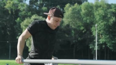 Güneşli sabahlarda paralel barlara triceps yapan şapkalı genç adam. Yaz aylarında şehir parkında sportif erkek eğitim yan görünümü. Adam barları egzersiz. 4k, Uhd. Orta atış