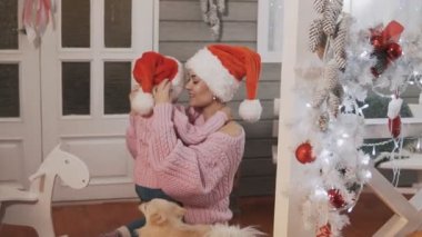 Genç anne ve kızı pembe pullovers ve kırmızı şapka Noel'de sarılma. Yavaş çekimde orta çekim. Mutlu anne Noel verandada onun çocuk kızı ile eğlenmek.