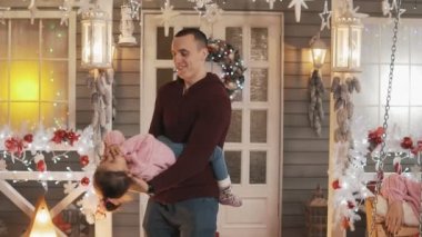 Mutlu baba Noel verandasında kızıyla eğleniyor. Babam küçük kızını yavaş çekimde gıdıklıyor. Noel günü pembe pullover baba ve kızı. Yakın çekim