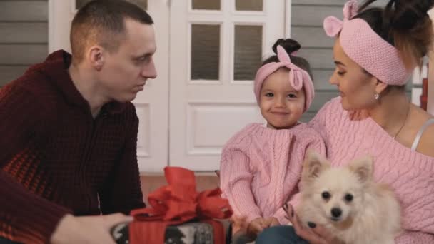 快乐的家庭包礼物为圣诞节 微笑的家庭与小女儿和狗坐在门廊与圣诞树 家庭概念 年轻的家庭在圣诞节拥抱 中等镜头 — 图库视频影像