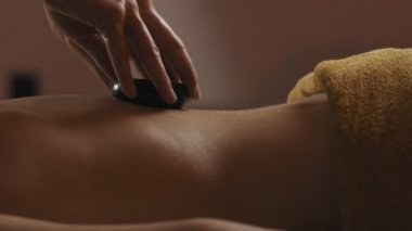 Spa salonunda sıcak taş masajı yaptıran genç kadın. Ağır çekimde masaj masasına yatan taşlar masajı sırasında dinlenen kadının yakın çekim. Akşam ışığı. 4k, Uhd