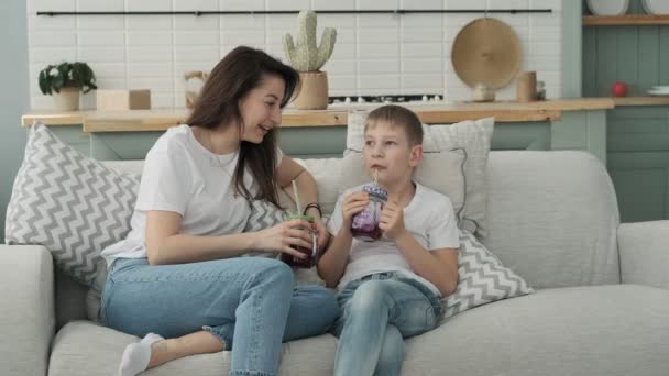 愛する母親は息子と一緒に時間を過ごす ママと息子は枕付きのソファに座って新鮮なジュースを飲みます 白いシャツと青いジーンズの家族 — ストック動画