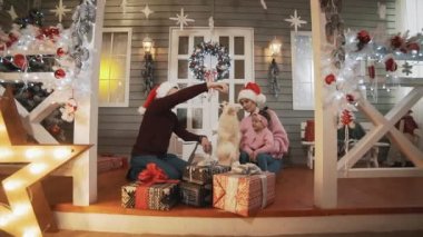 Küçük kızları ve köpekleri olan genç bir aile Noel ağacı ve dekorla verandada oturuyor. Yavaş çekimde mutlu aile kavramı. Kazaklı genç bir aile Noel günü sarılıyor.