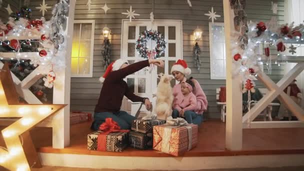 小さな娘と犬がクリスマスツリーと装飾とポーチに座っている若い家族 スローモーションで幸せな家族の概念 若いです家族でプルオーバー抱擁でクリスマスの日 — ストック動画