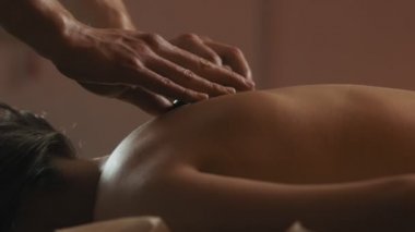Sıcak taş terapisi sırasında spa merkezinde dinlenen genç beyaz kadın sıcak akşam ışığı ile. Salonda masaj masasında yatan bir kadının yakın çekim. 4k, Uhd