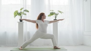 Hamile kızıl saçlı kadın yoga yapıyor. İçeride savaşçı pozisyonunda duruyor.