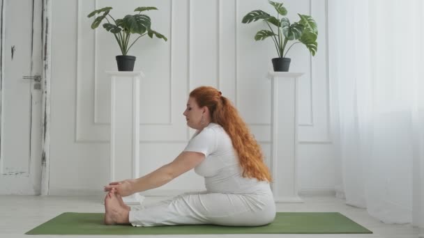 Hermosa madre practicando yoga en un apartamento blanco sentado en una esterilla verde en UHD — Vídeos de Stock