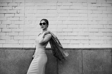 Fashion street style portrait of swag girl against wall. Wearing jeans jacket, casual dress and sunglasses. Trendy outdoor lifestyle and stylish urban fashion concept. Black and white photography.