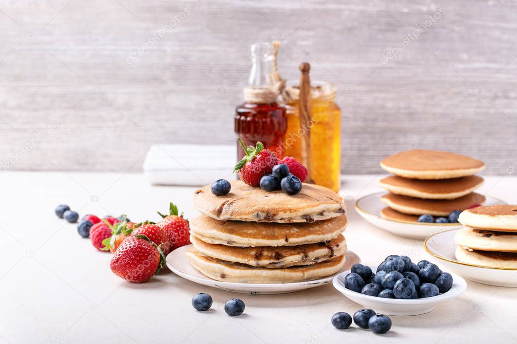 Blueberry pancakes served with strawberries, blueberries, raspberries, honey and maple syrup over white background