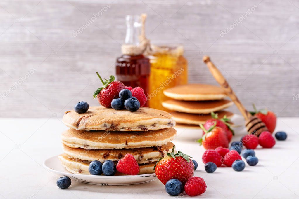 Blueberry pancakes served with strawberries, blueberries, raspberries, honey and maple syrup over white background