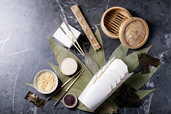 Noodles and rice on bamboo leaves with traditional chinese takeaway boxes, wooden steamer and chopsticks over dark texture backround. Top view, flat lay. Copy space