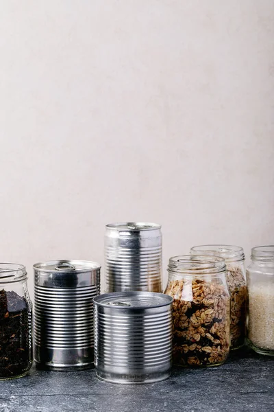 Canned food in metal cans with oats, rice, tea, and pasta in glass jars over white texture background. Copy space. Food supply concept